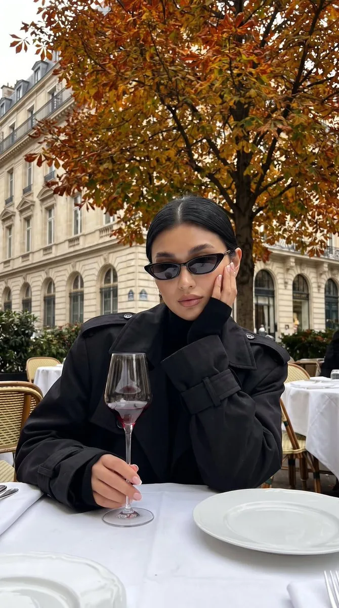 A woman in a black trench coat sitting at an outdoor cafe with wine.