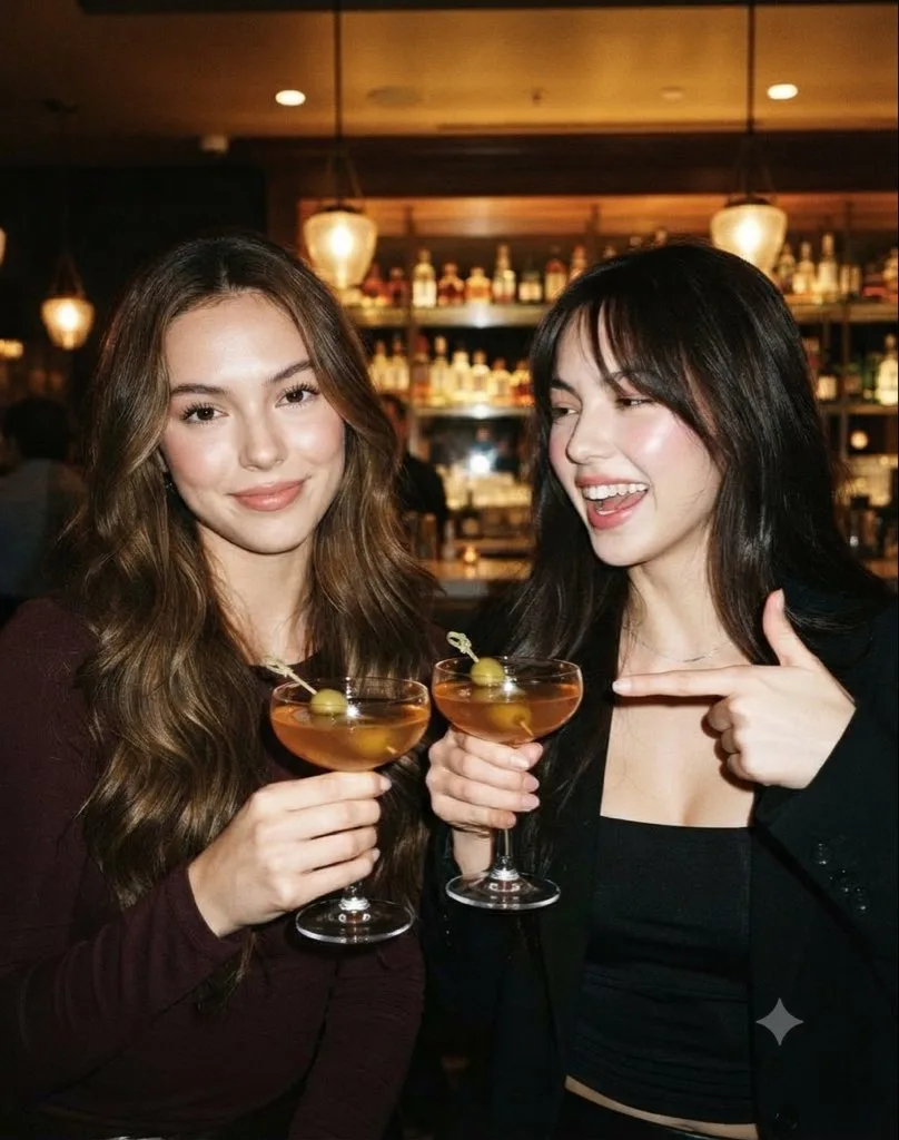 Two women laughing while holding cocktails at a dimly lit, upscale bar.