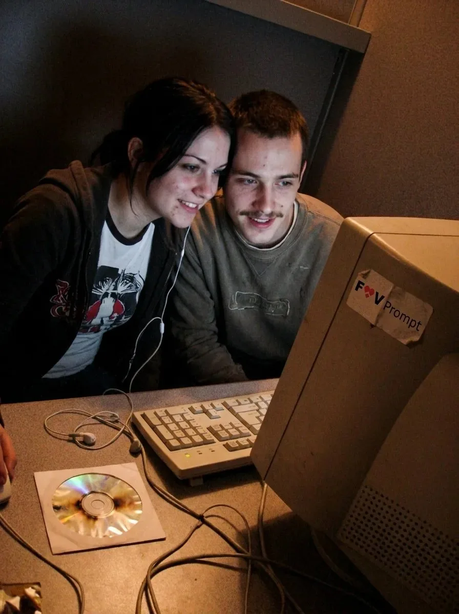 A candid low-resolution 2000s webcam photo of a young couple chatting in an internet cafe.