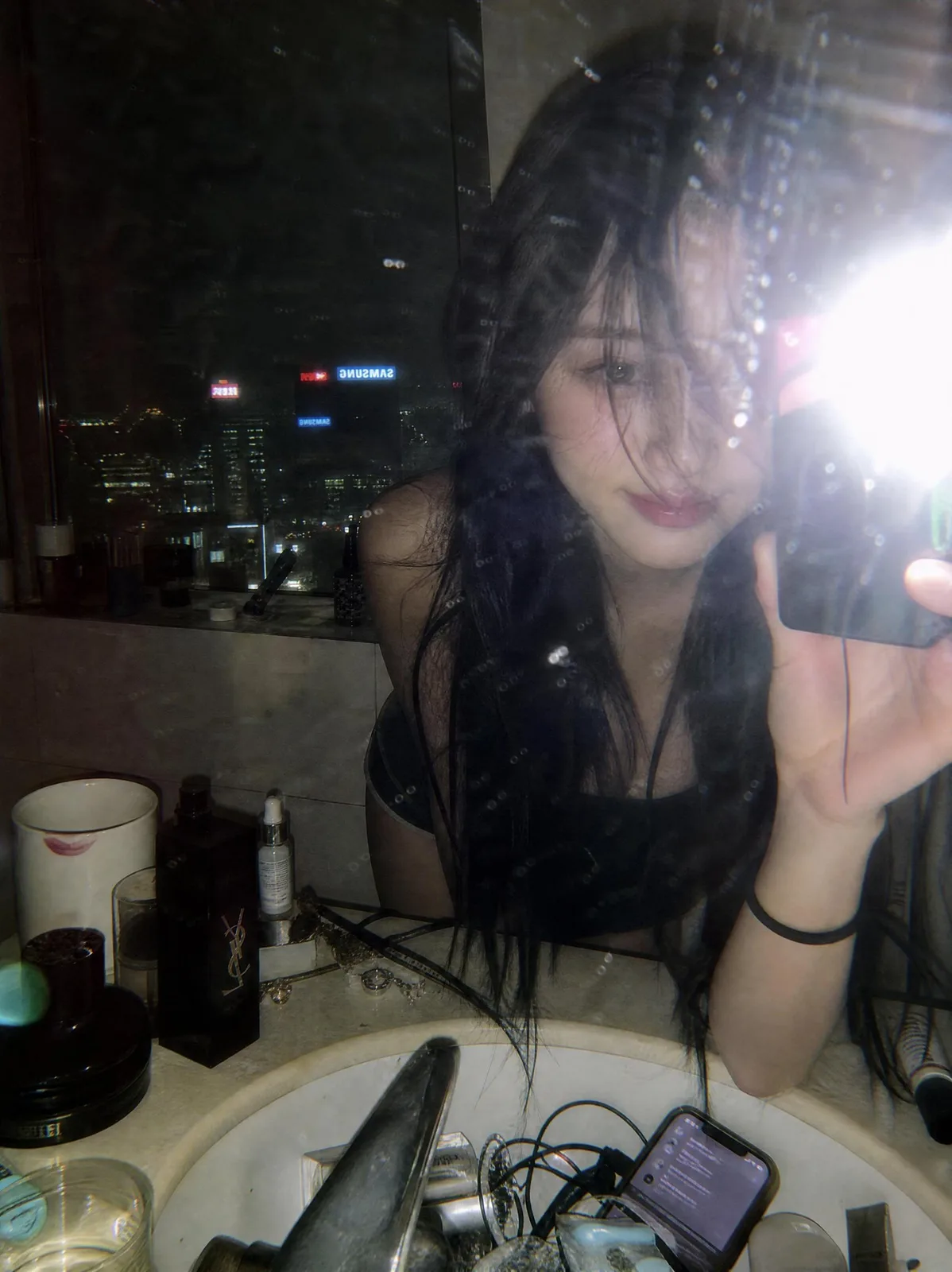 A candid mirror selfie of a woman in a luxury bathroom with city lights in the background.