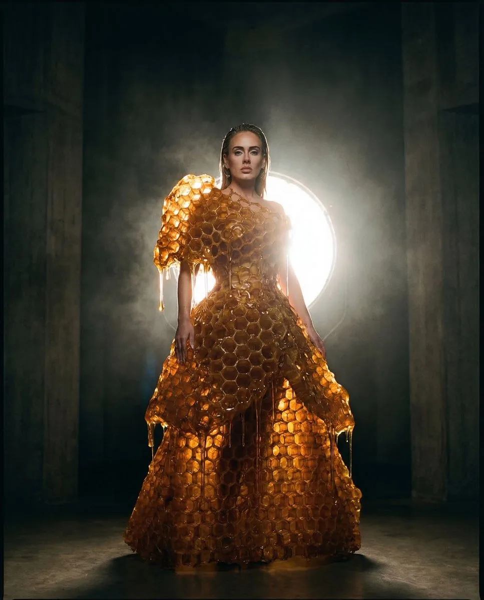 A minimalist figure in a honeycombed amber gown within a dark concrete chamber, heavily backlit. (2)