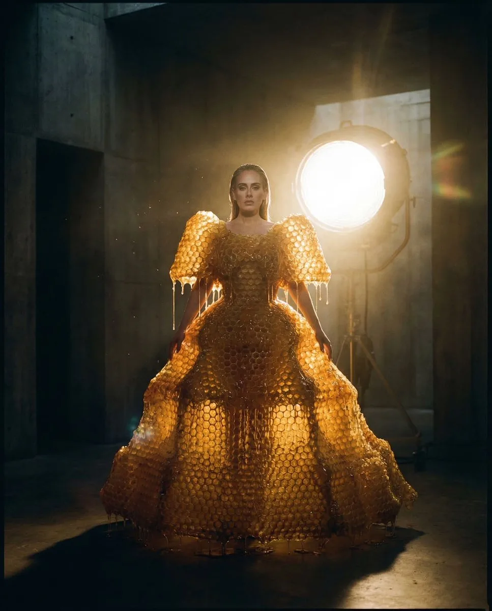 A minimalist figure in a honeycombed amber gown within a dark concrete chamber, heavily backlit. (3)